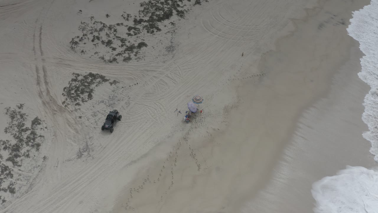 Overhead frame from Los Cabos, Mexico showing a black buggy near beach umbrellas on pale sand, with scrub patches, tire tracks, white surf at the right edge, and no visible people.