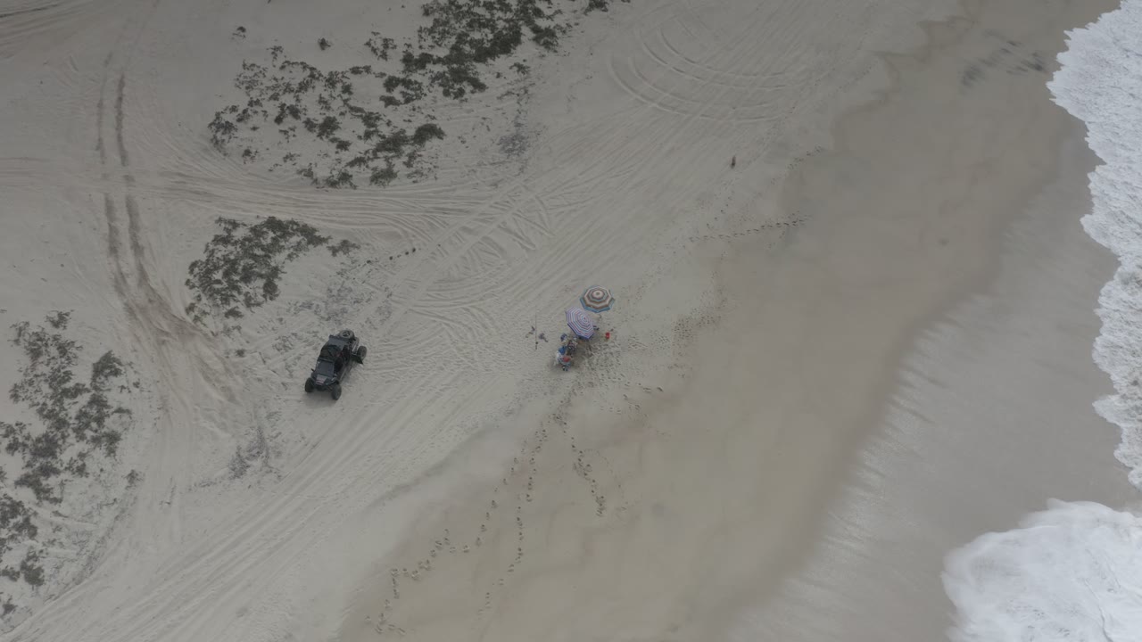 High overhead Los Cabos beach view with the black buggy left of center, two umbrellas on the sand, faint tire arcs, scrub clusters, and a band of white surf, with no visible people.