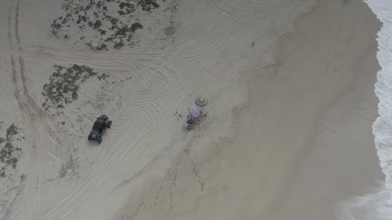 Top-down sandy beach with buggy, umbrellas, tire marks, scrub, and surf.