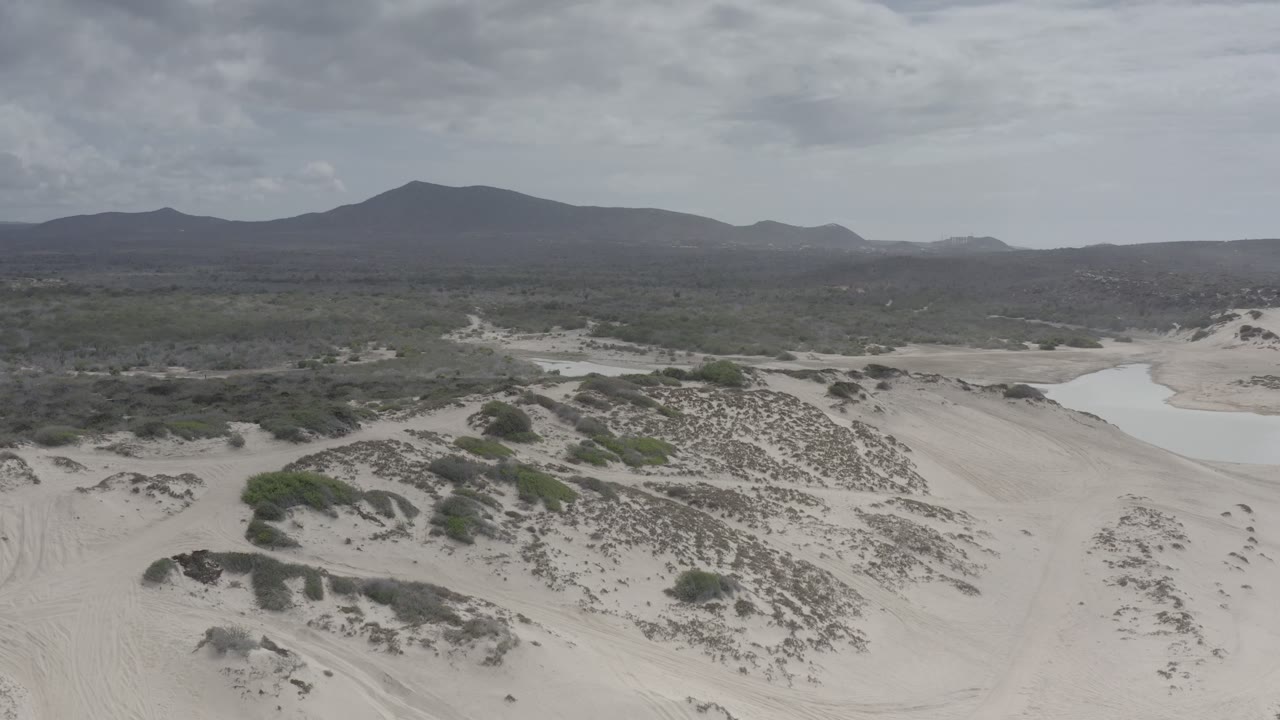 A Los Cabos aerial frame looks across sandy dune ridges and low vegetation toward a shallow water pocket, dark mountain ridge, overcast sky, and no visible people.