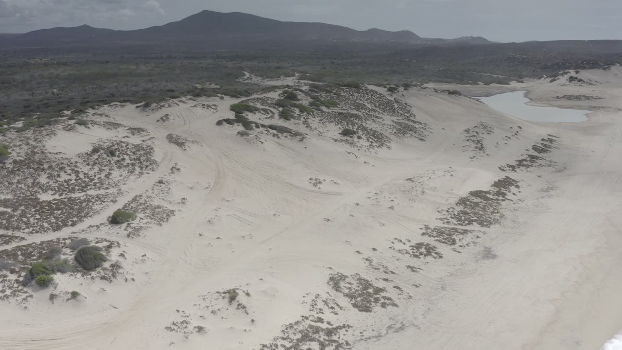 Aerial frame of Los Cabos sand dunes and scrub with a pale water channel near the upper right, distant hills across the horizon, and no visible people.