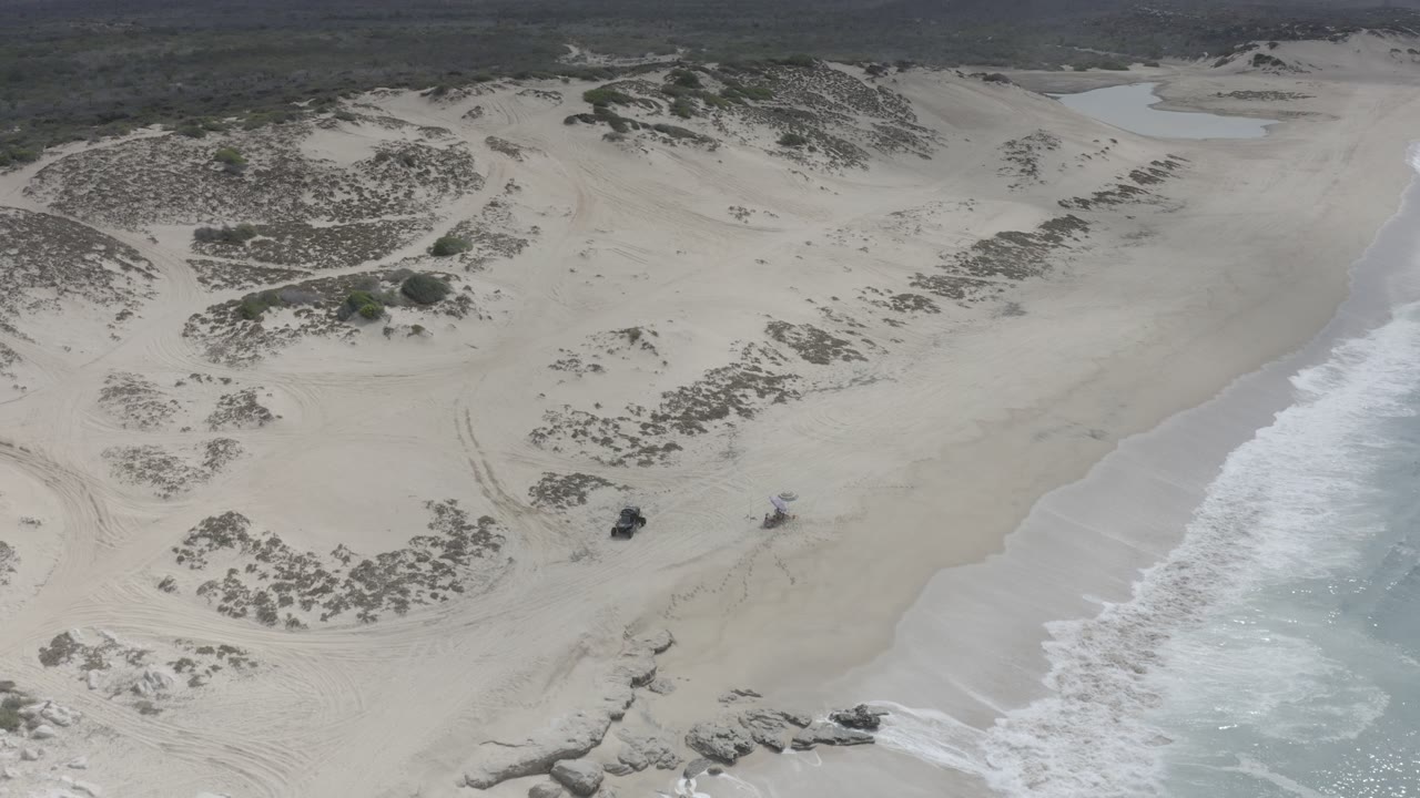 Oblique aerial view of a gray sandy Los Cabos beach with scrub-covered dunes, dark rocks near the lower edge, white surf on the right, and no visible people.