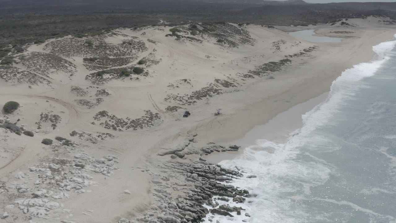 Aerial view of surf breaking beside a sandy beach and dark shoreline rocks.