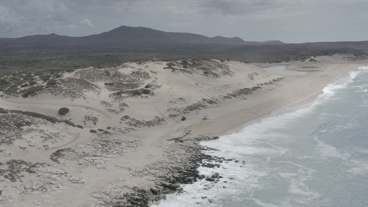 Aerial view over Los Cabos, Mexico shoreline with foamy surf at right, pale sand and dunes across the center, dark rocks in the foreground, distant hills under cloudy sky, and no visible people.