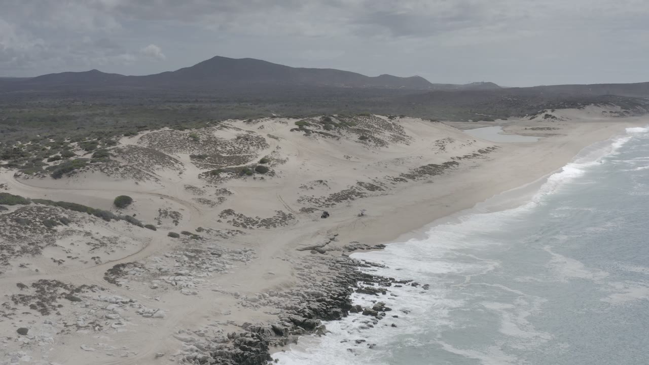 Wide aerial coastline with pale dunes, surf, rocks, and hills in the background.