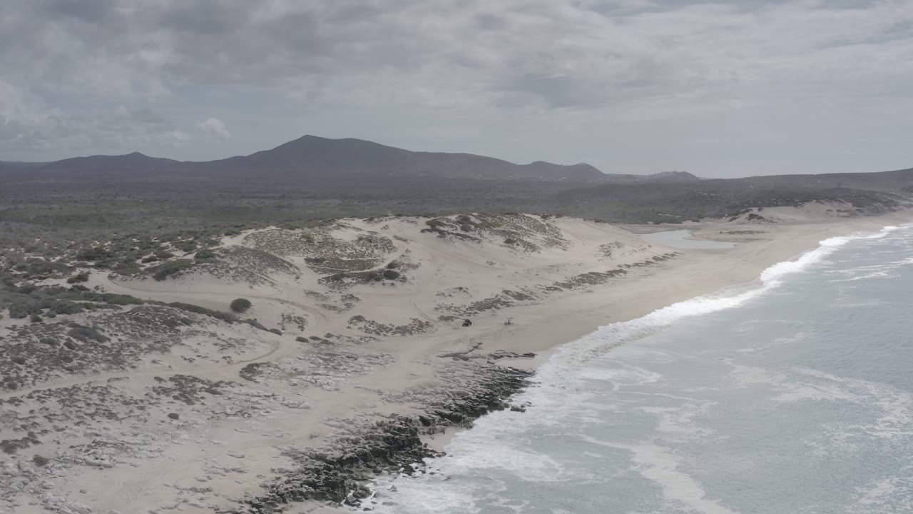 Aerial coastline with sand dunes, surf, rocky shore, and distant mountains.