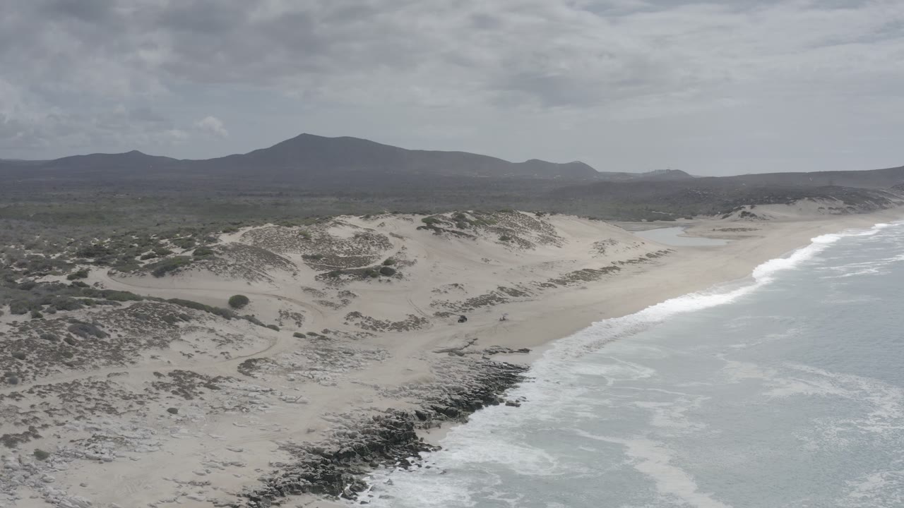 Aerial frame over Los Cabos showing the beach curving beside pale blue-green water, dune ridges, scattered scrub, mountain silhouettes, and no visible people.