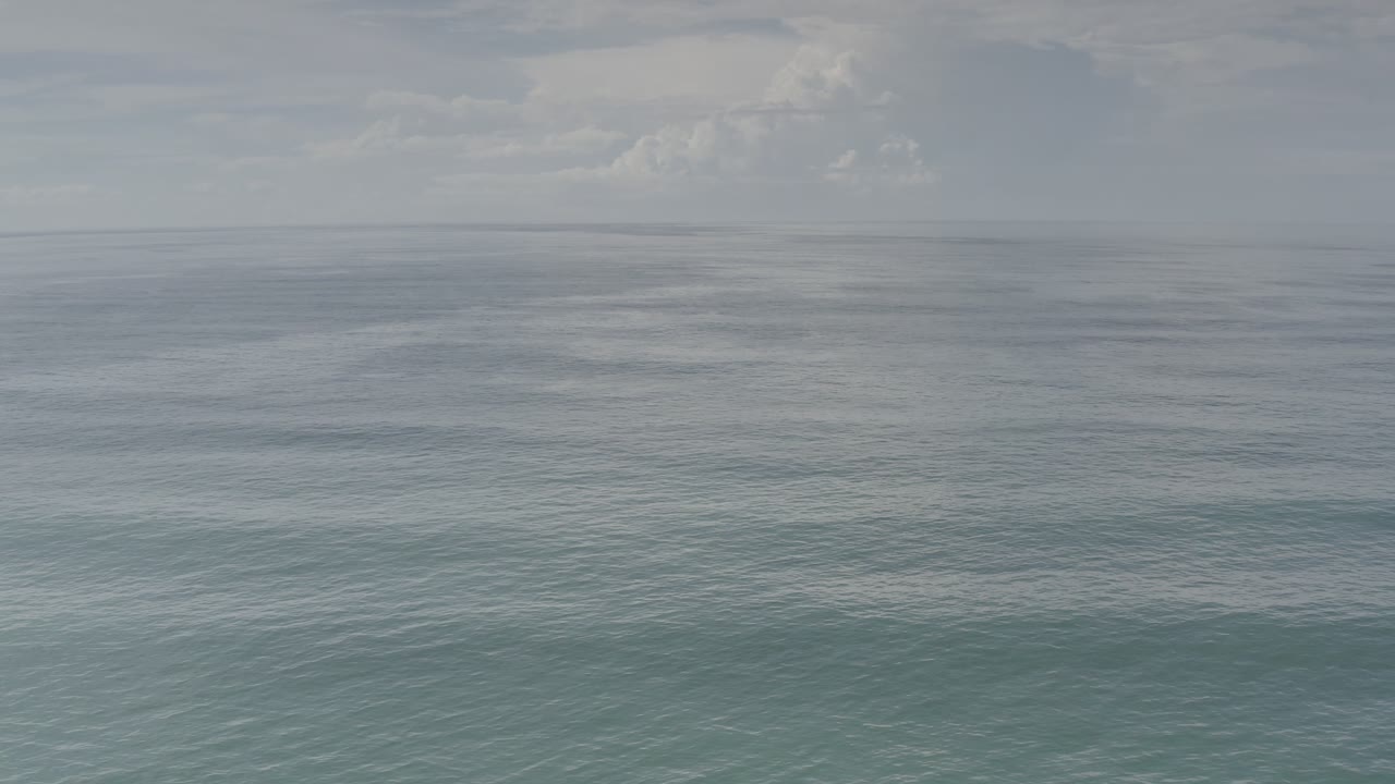 Open Pacific water in Los Cabos, Mexico, with pale turquoise foreground tones, darker water near the horizon, low cloud bands, and no visible people.