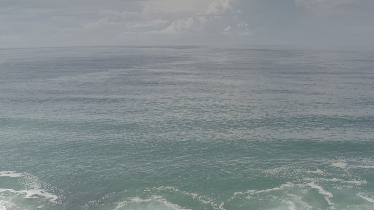 Opening aerial frame of the Los Cabos, Mexico coastline with gray-blue open water, white surf near the lower edge, a cloudy horizon, and no visible people.