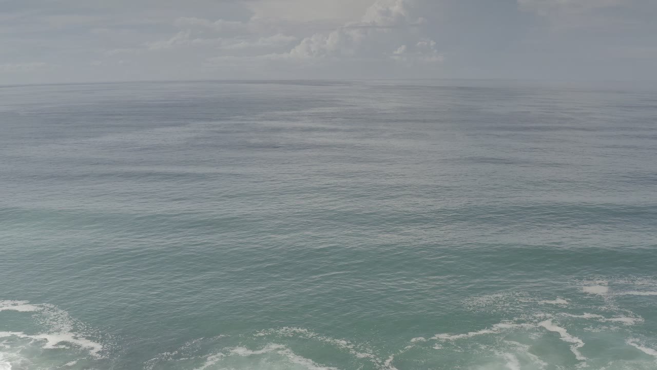 Los Cabos aerial coastline frame with teal water bands, white breaking waves across the lower portion, overcast sky, and no visible people.