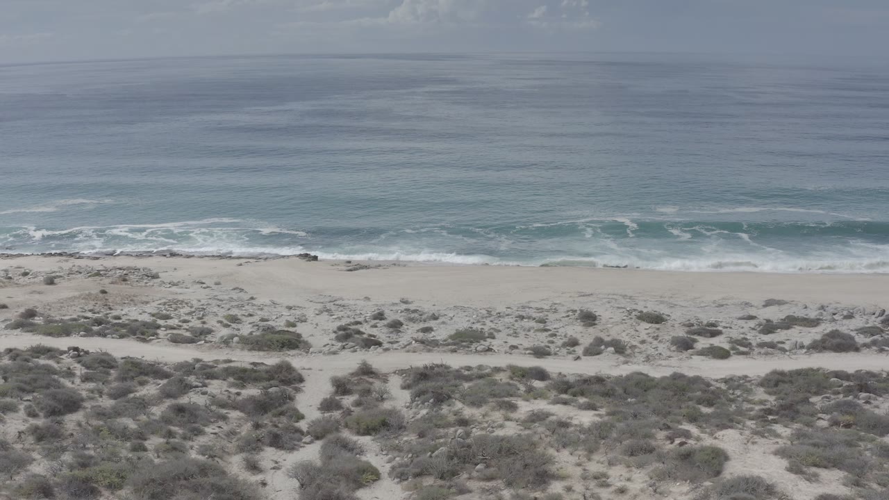 Aerial frame over Los Cabos, Mexico, showing scrub dunes in the foreground, a pale beach, blue-green ocean, and no visible people.
