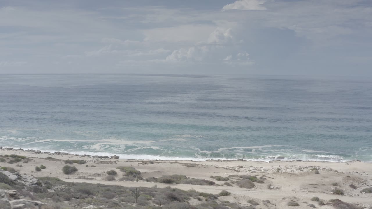 Elevated Los Cabos coastline view with pale sand, low scrub, white surf, blue-green ocean, soft clouds, and no visible people at the start of the clip.