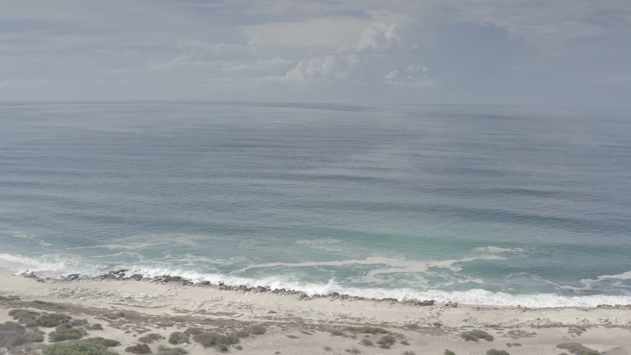 Aerial view over the Los Cabos shoreline, with blue-gray open water, white surf, pale sand, scattered rocks, and no visible people.