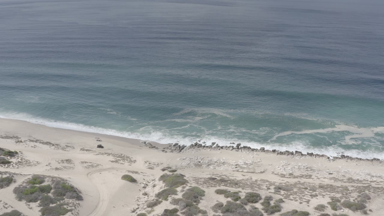 Aerial view over a Los Cabos, Mexico beach with blue-green water, white surf, pale sand, dune scrub, and no visible people at the start of the clip.
