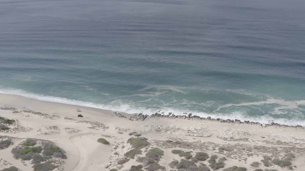 Overhead Los Cabos shoreline where a band of white surf breaks onto pale sand, with low scrub and vehicle tracks visible inland and no visible people.