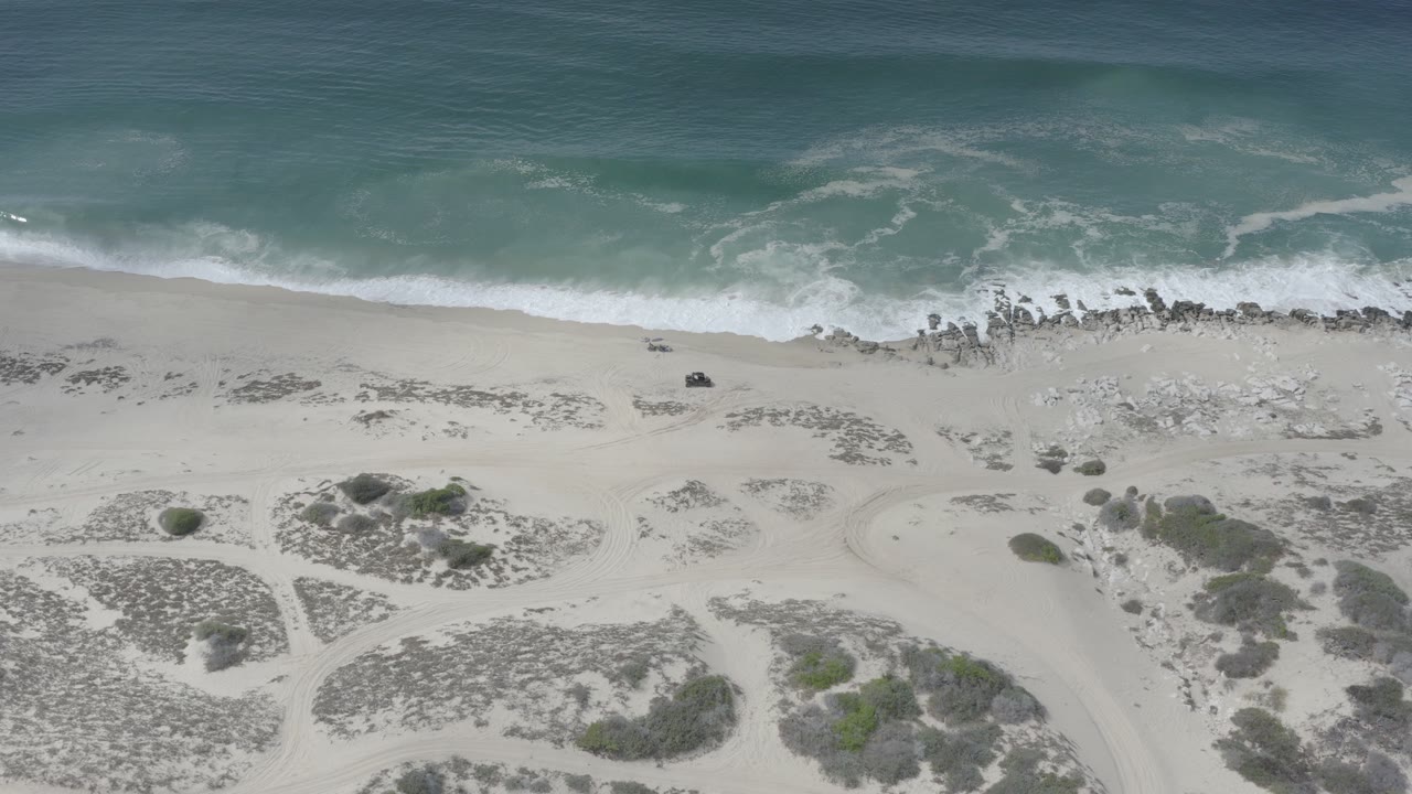 Overhead view in Los Cabos, Mexico, showing teal ocean surf reaching pale sand, scattered dark rocks near the waterline, dune vegetation below, and no visible people.