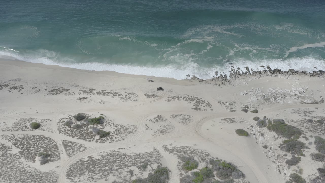 Drone-like overhead view of surf, sand, rocky waterline, and scrub dunes.