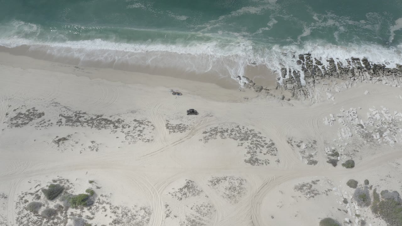Overhead beach with surf, rocks, sand, scrub, and tire tracks.