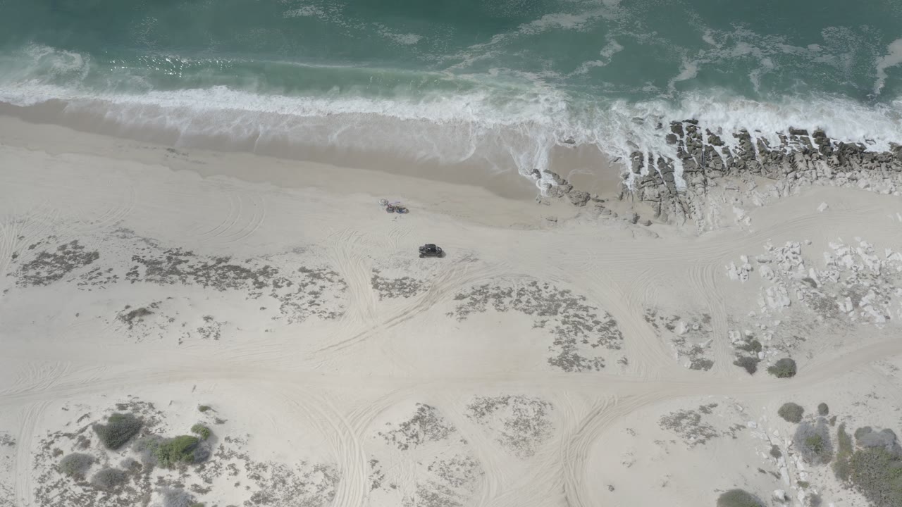 A Los Cabos aerial frame shows white foam spreading across dark shoreline rocks, with pale sand, curved tire marks, scrub clusters, and no visible people.