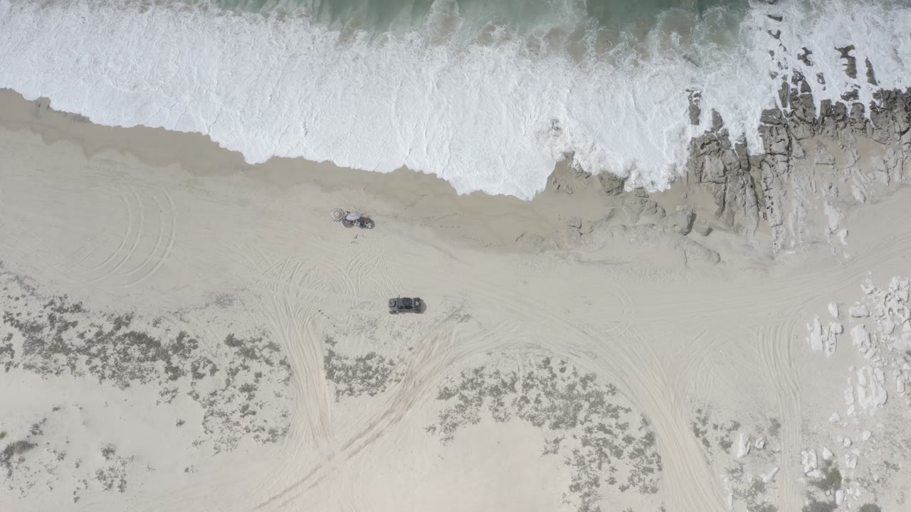 Overhead view of a Los Cabos beach with white surf along the top edge, pale sand, dark rocks, faint tire tracks, and no visible people.