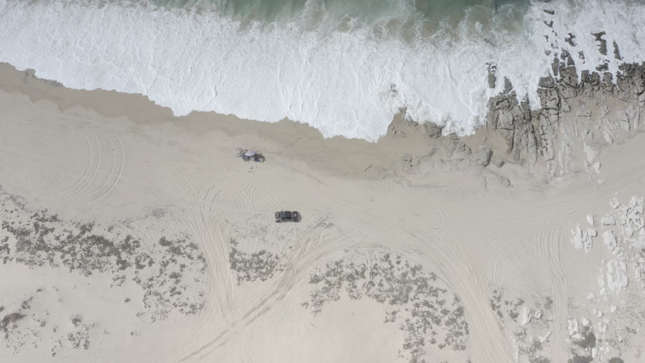 Aerial Los Cabos beach frame with foamy water pushing over rock outcrops, a wide sandy area, small dark beach objects, and no visible people.