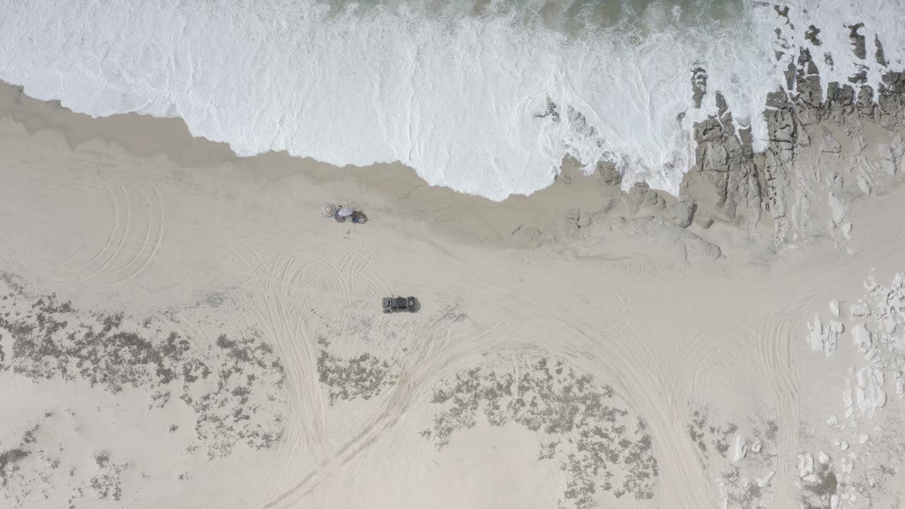 Top-down beach shoreline with surf, rocks, sand, and faint vehicle tracks