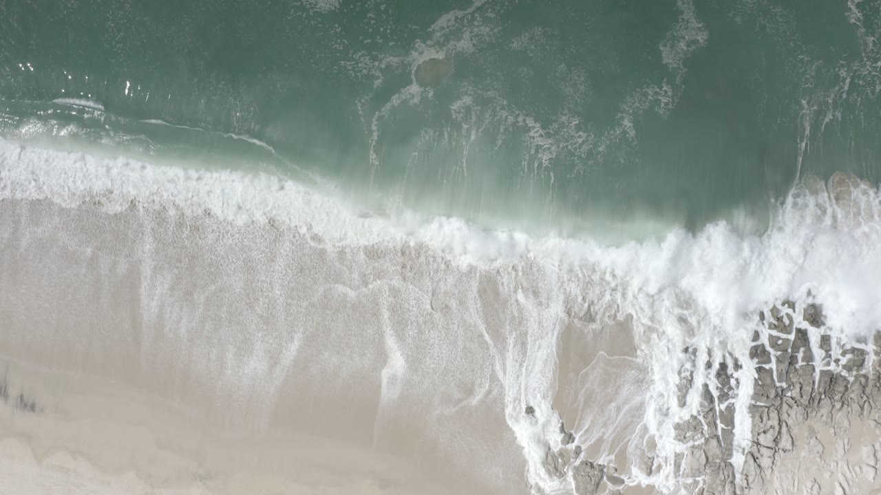 Overhead view of turquoise water and white surf washing onto pale sand in Los Cabos, Mexico, with no visible people.