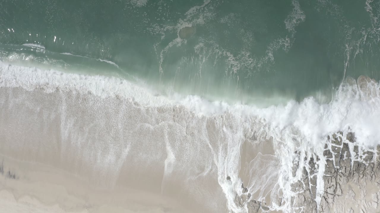 White wave foam spreads across the pale beach below turquoise water in Los Cabos, Mexico, with no visible people.