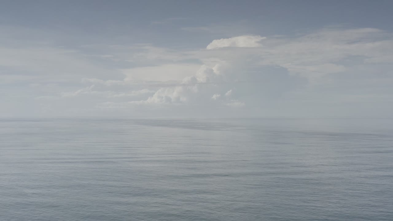 Los Cabos ocean view one second in, with muted water texture across the frame, pale haze at the horizon, distant white clouds, and no visible people.