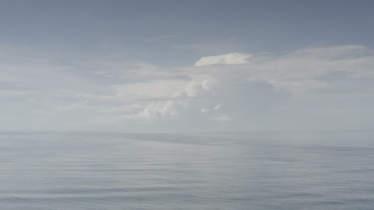 Opening frame of a pale blue sky over the ocean in Los Cabos, Mexico, with a broad white cloud mass above the horizon and no visible people.