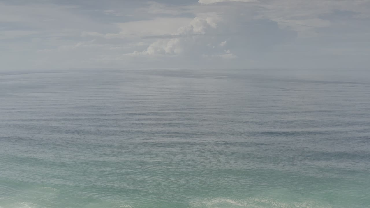 Early frame over the Los Cabos sea showing smooth gray water, a faint turquoise band near shore-facing water, soft clouds, and no visible people.