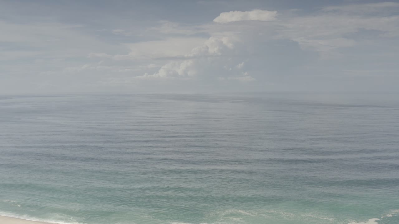 Wide Los Cabos ocean view at the start of the clip, with gray-blue water, turquoise shallows, soft clouds, and no visible people.