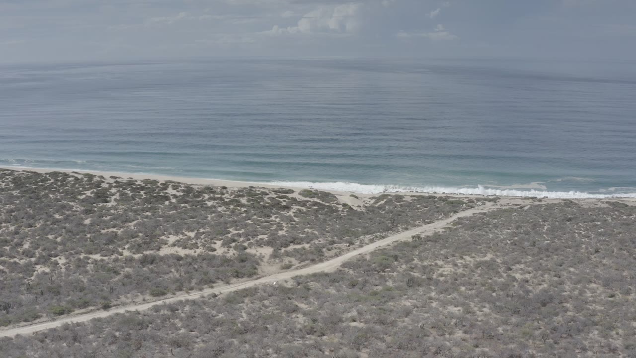 Aerial view in Los Cabos, Mexico, showing a winding dirt track through low brush below a thin beach and calm blue ocean, with no visible people.