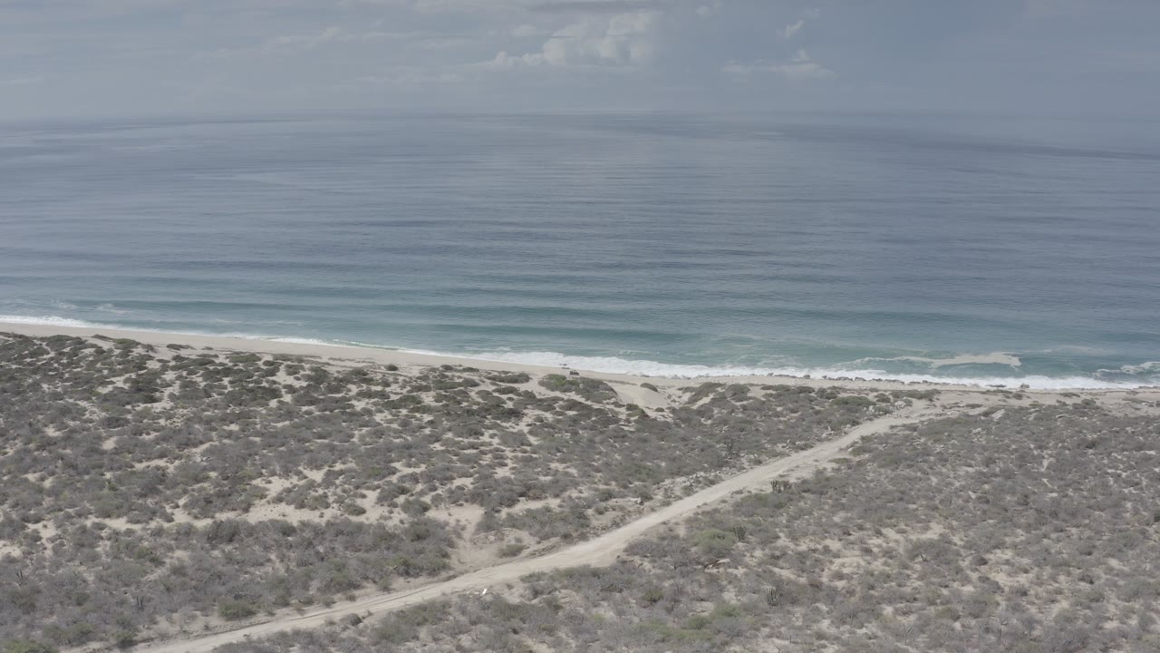 Opening aerial frame over the Los Cabos coast, with scrubland and a pale dirt road below, a narrow beach, blue water, and no visible people.