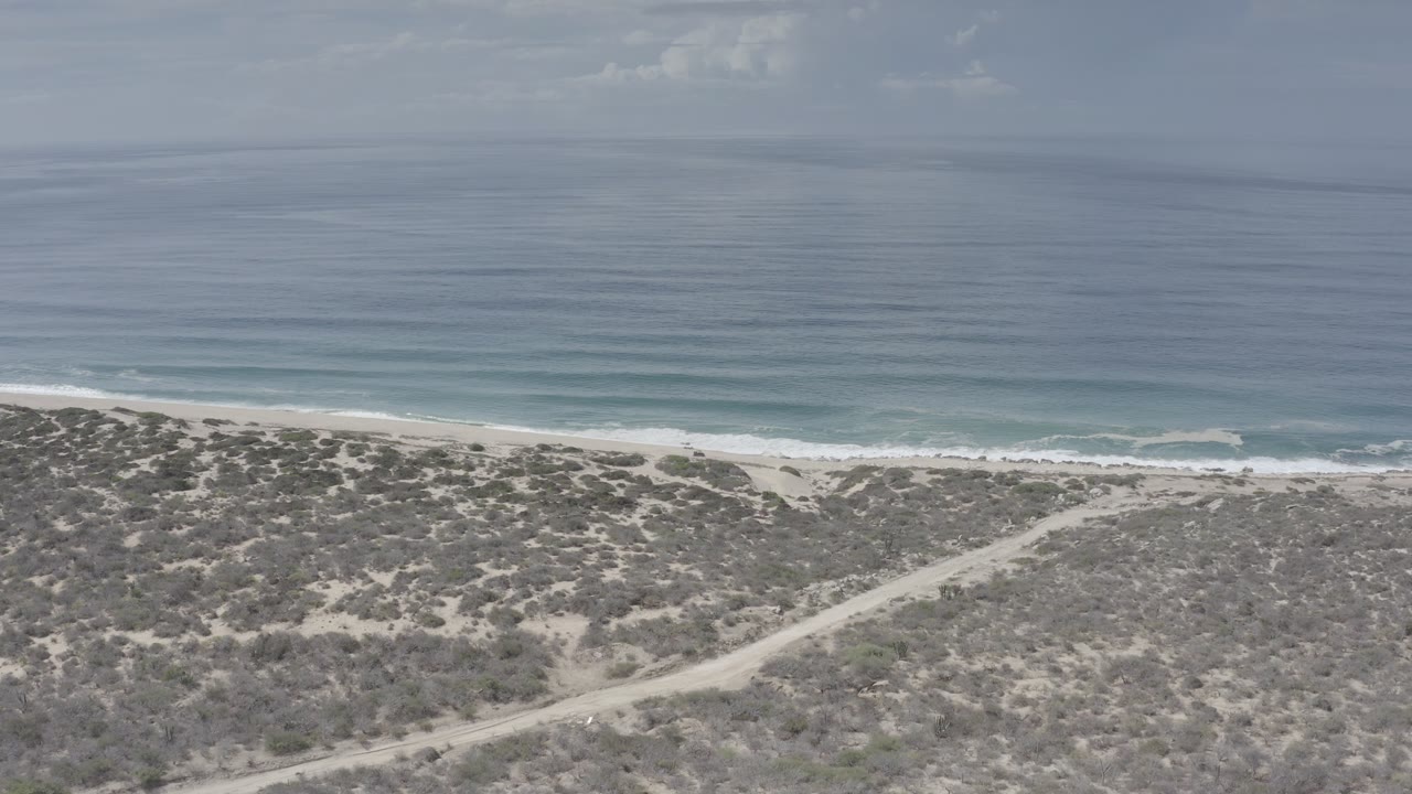 Early frame from the Los Cabos aerial view, showing the dirt track angling through scrub toward the beach while small waves line the shore and no visible people appear.