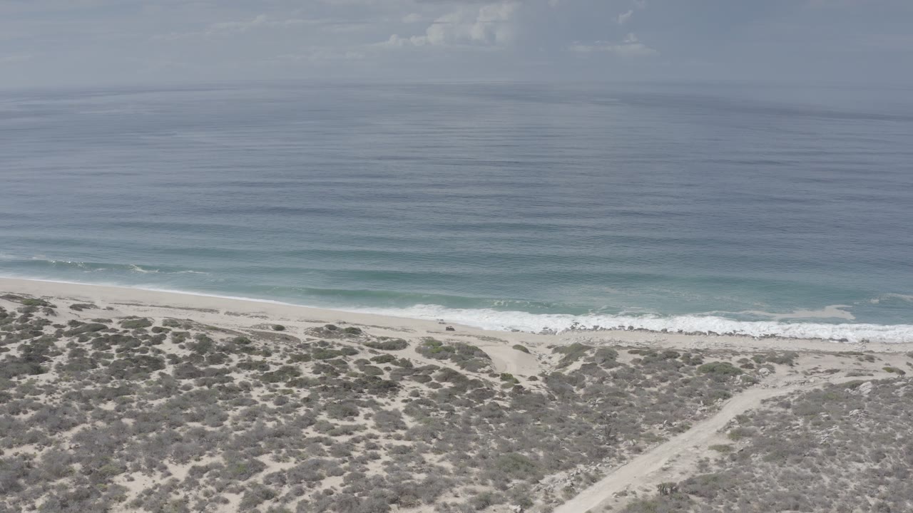 Opening aerial frame over a Los Cabos shoreline with gray-blue water, pale sand, scrubby dunes, and no visible people.