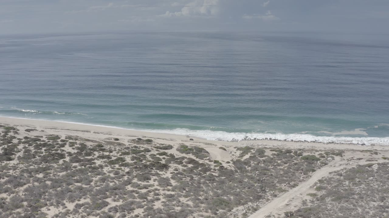 Wide coastal view with waves, beach, dunes, and gray sky.