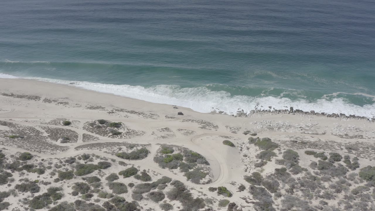 Aerial view in Los Cabos, Mexico with pale sand dunes, scattered scrub, a white surf line, turquoise water, and no visible people at the start of the clip.