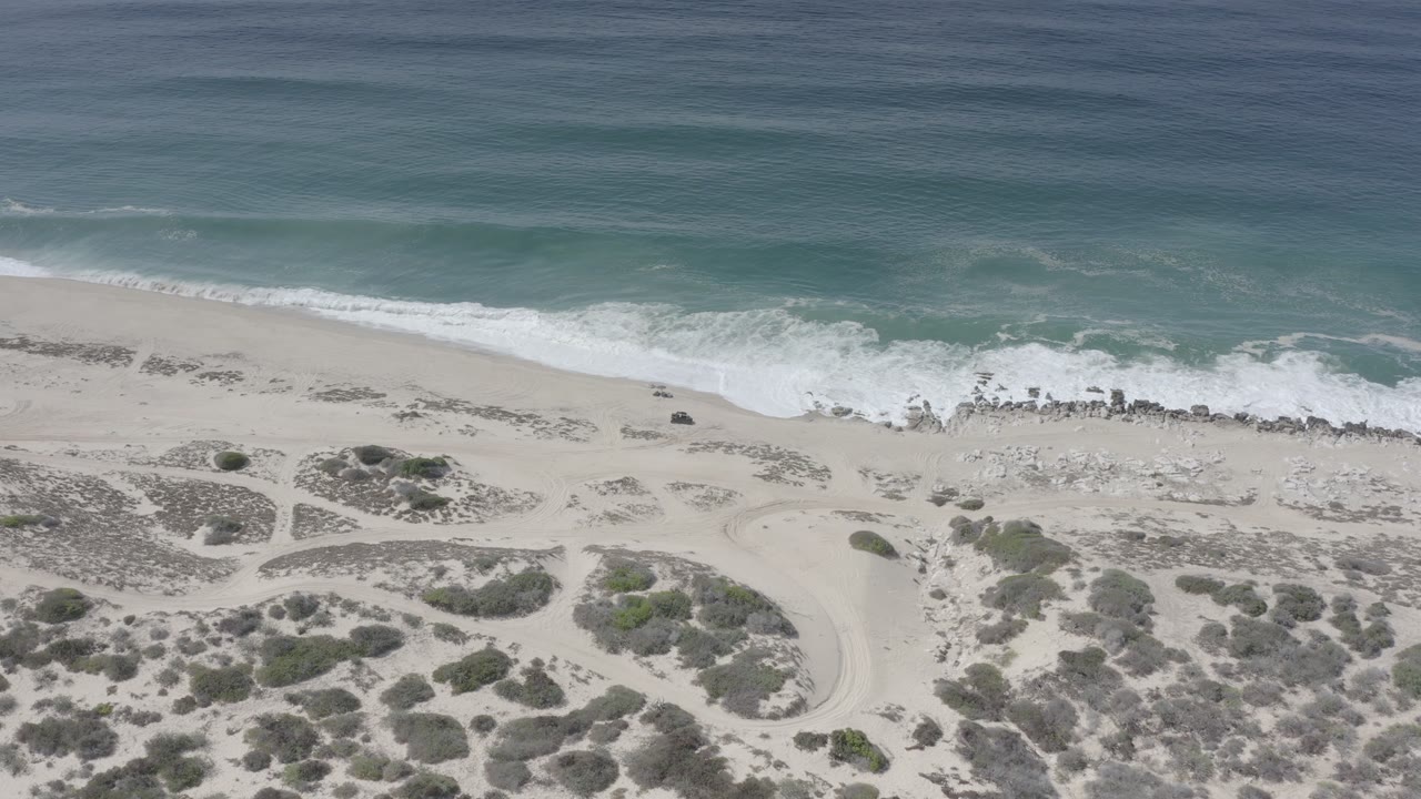 Drone-like view of empty sandy coast, scrub, surf wash, and blue-green water.