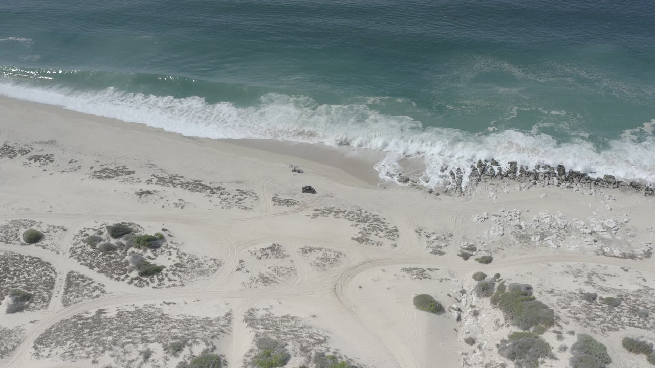 Overhead view of a Los Cabos beach with teal water, white surf, pale sand, scattered rocks, dune scrub, and no visible people at the start of the clip.