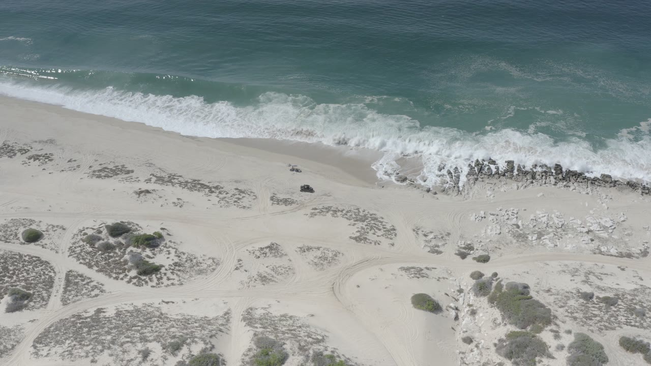 Aerial Los Cabos shoreline frame showing a band of foamy surf reaching the sand, darker rocks near the waterline, scrub patches inland, and no visible people.