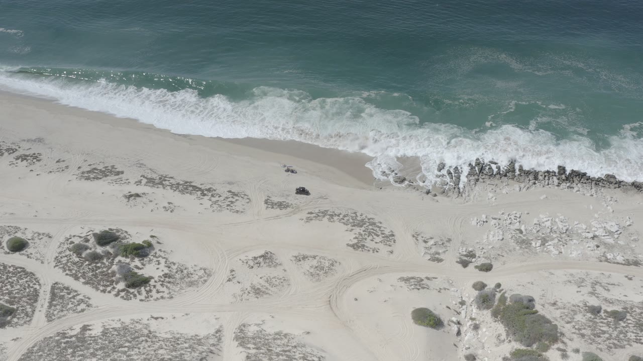 Drone view of waves, rock clusters, sand, and low beach vegetation.