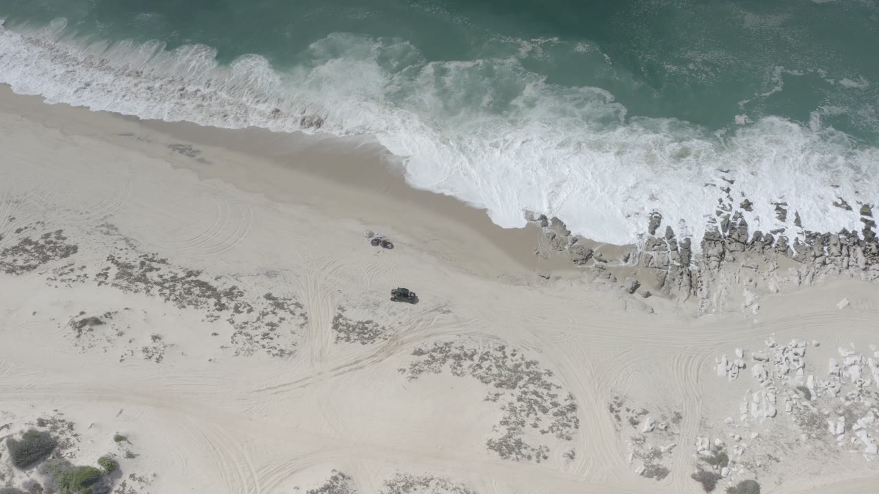 Overhead view of a Los Cabos, Mexico beach where white surf reaches pale sand beside exposed rocks, with two dark vehicles or objects on the beach and no visible people.