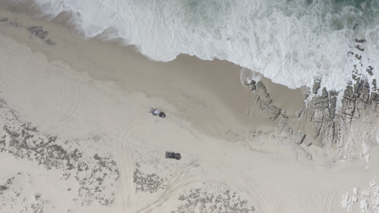 Overhead Los Cabos beach frame showing the wave edge spread across the top of the sand, rocks near center right, and no visible people.