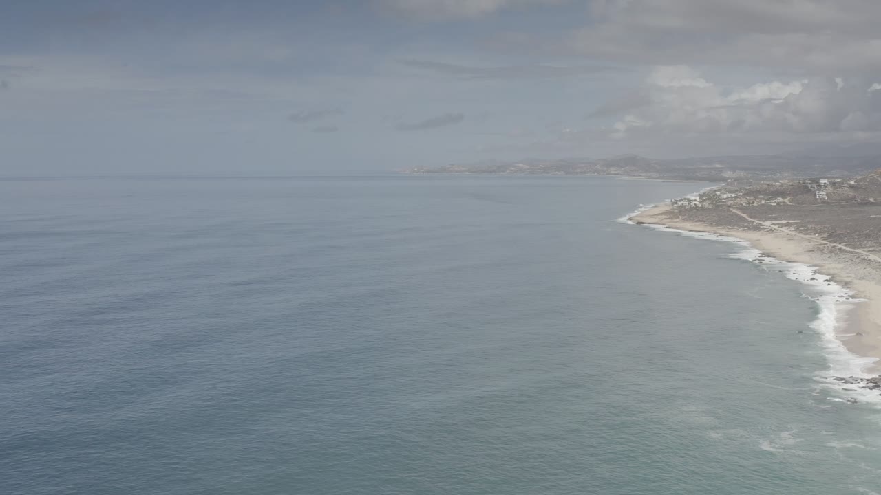 Wide aerial coastline with ocean, surf, sandy shore, and cloudy sky.