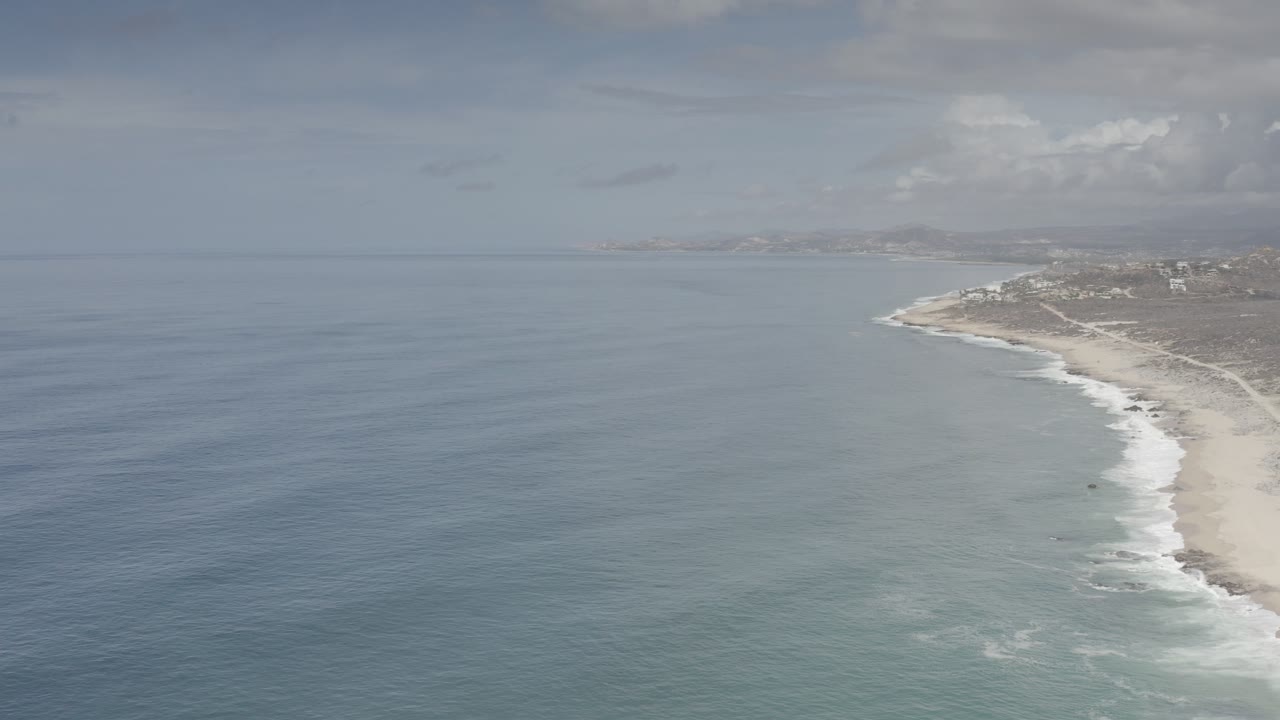 High aerial view over the Los Cabos coastline with blue-green water on the left, white surf on the beach to the right, distant shore buildings, and no visible people.
