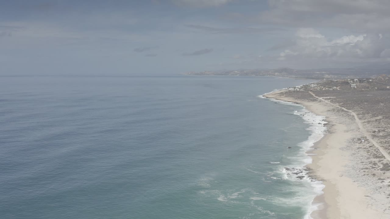 High aerial frame of the Los Cabos shore with the ocean filling the left side, foam lines along the sand, dunes on the right, and no visible people.