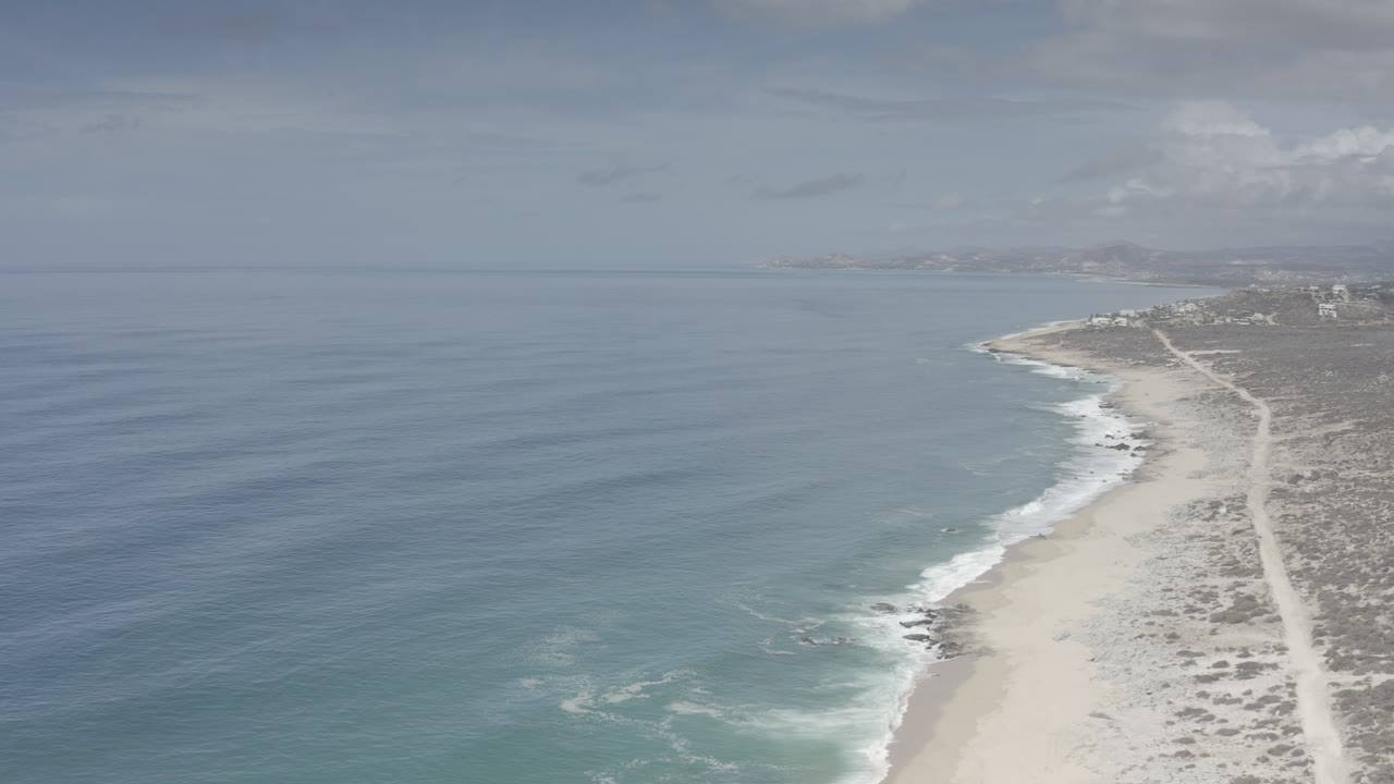 High aerial view of the Los Cabos coastline with blue ocean on the left, sandy beach on the right, a coastal track inland, and no visible people.
