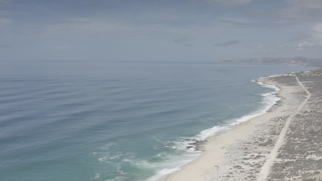 Aerial view of blue ocean beside a pale beach and scrub dunes.