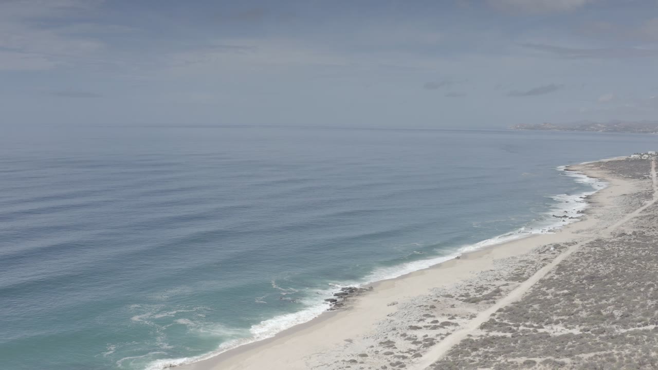 Los Cabos aerial frame with calm blue water spreading across the left side, a pale beach line at lower right, scrubby terrain inland, and no visible people.